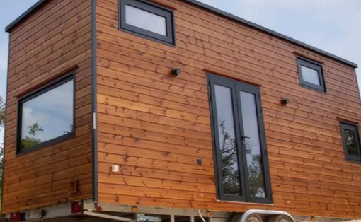 Wood-clad tiny house on a trailer, raised on wheels, with large black-framed glass doors and several square windows on the side.
