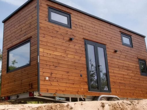 Wood-clad tiny house on a trailer, raised on wheels, with large black-framed glass doors and several square windows on the side.
