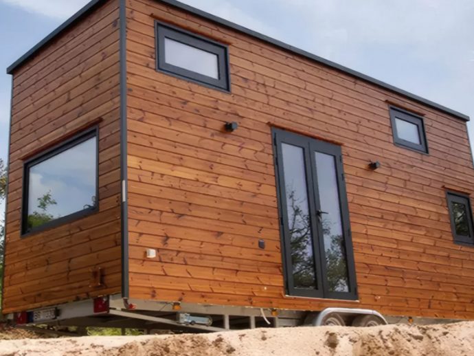 Wood-clad tiny house on a trailer, raised on wheels, with large black-framed glass doors and several square windows on the side.