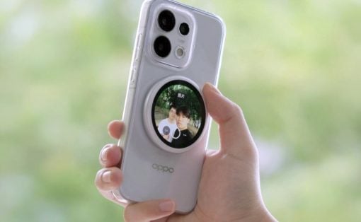 Hand holding a light gray OPPO smartphone with a circular photo display on the back, outdoors with a green blurred background.