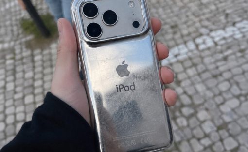 Hand holding a shiny silver iPod with Apple logo and 'iPod' text in a clear case, outdoors on a cobblestone pavement.