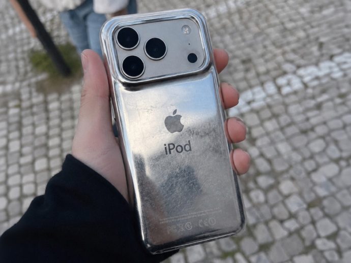 Hand holding a shiny silver iPod with Apple logo and 'iPod' text in a clear case, outdoors on a cobblestone pavement.